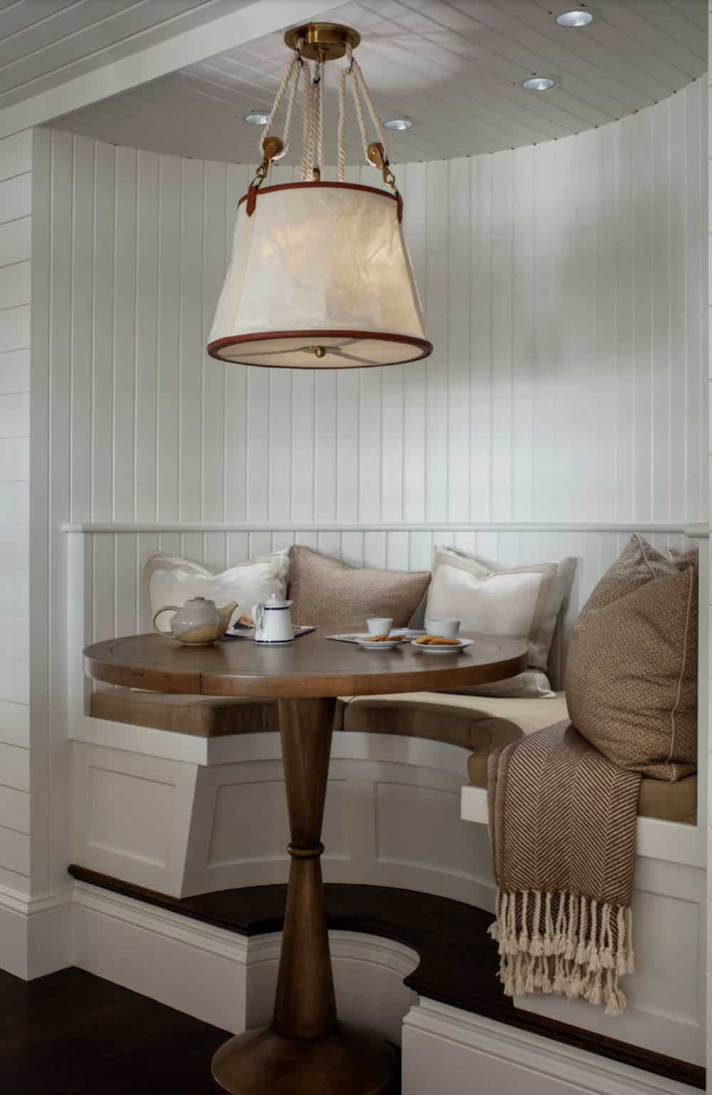 Cozy built-in banquette nook with curved white beadboard walls, round pedestal table, linen cushions, and nautical rope pendant light