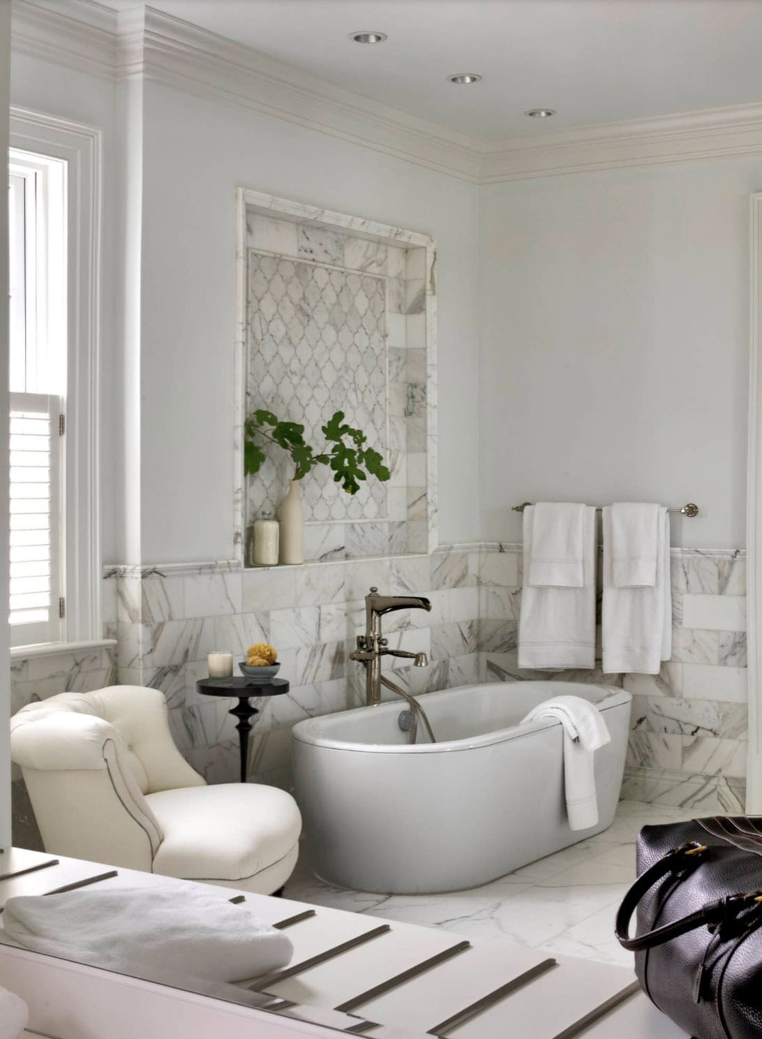 Bright all-white bathroom with freestanding soaking tub, marble lattice tile niche, polished nickel faucet, and tufted lounge chair