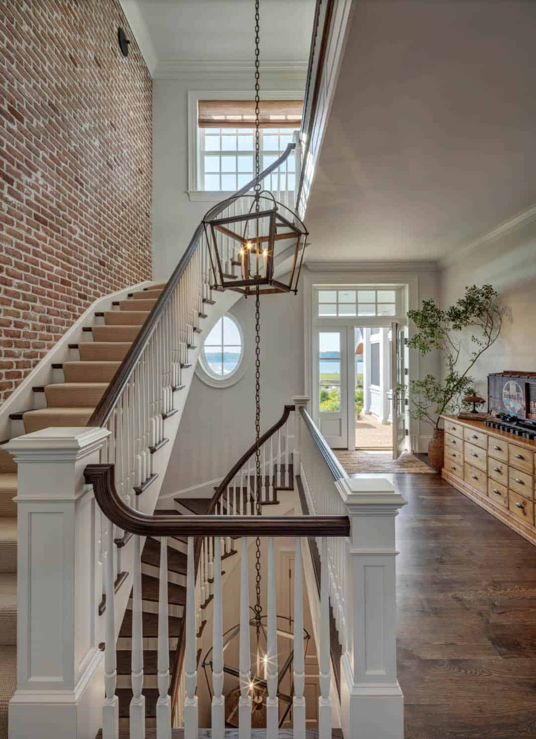 Two-story stair hall with curved staircase, exposed brick accent wall, large lantern pendant, oval porthole window, and front door open to water view