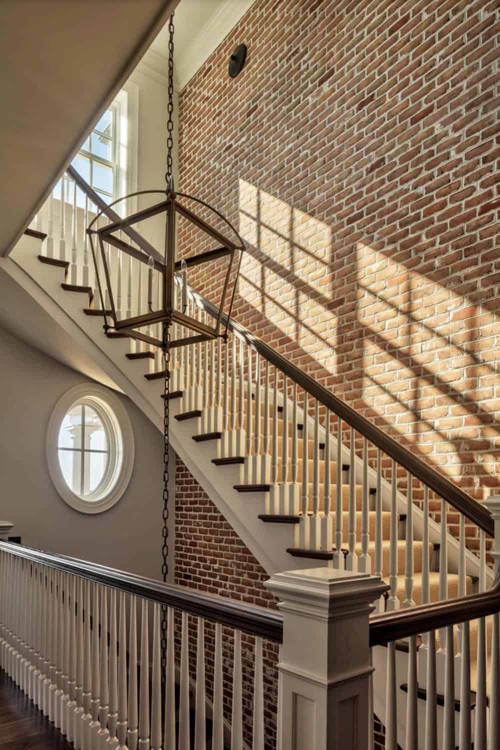 Close-up of curved staircase against sunlit exposed brick wall with oval porthole window and hanging bronze lantern pendant