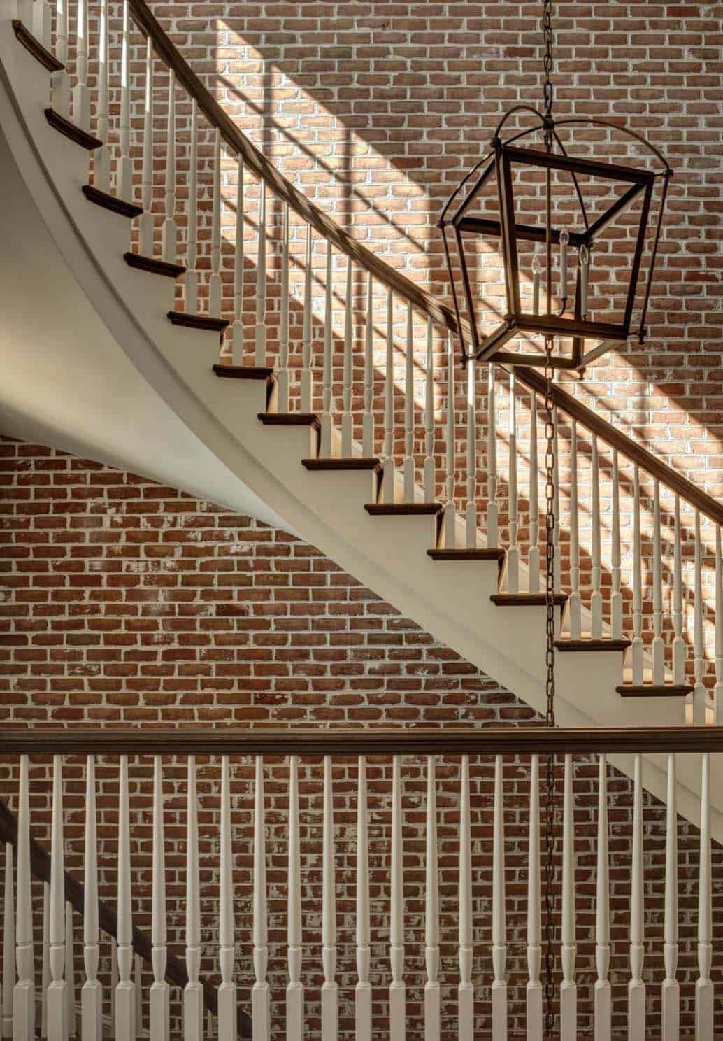 Detail of sweeping curved staircase with white balusters, dark wood handrail, and hanging iron lantern against aged brick wall