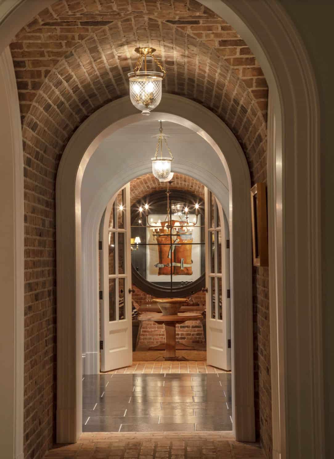Brick-vaulted barrel corridor leading through arched double doors to a candlelit wine cellar room beyond