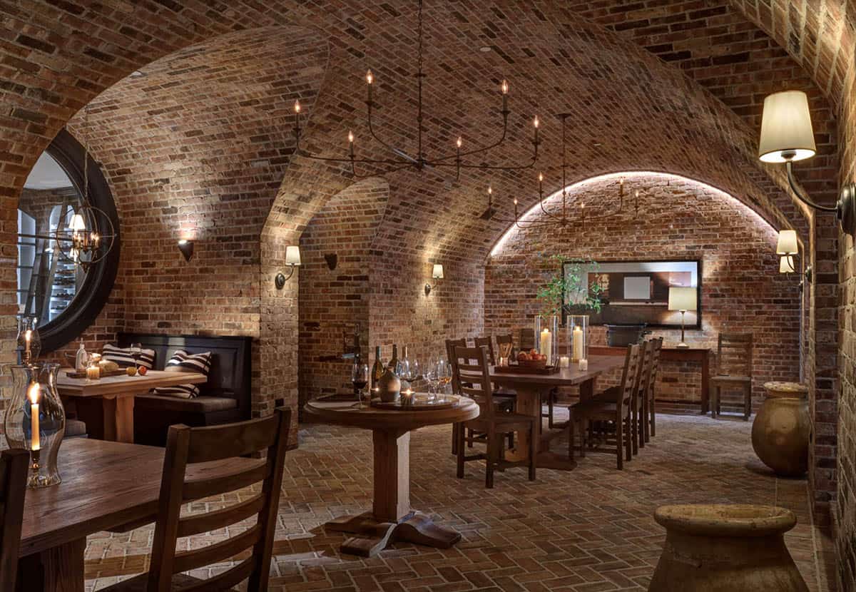 Underground wine cellar dining room with soaring brick barrel-vaulted ceiling, iron chandeliers, rustic wood tables, and ambient candlelight