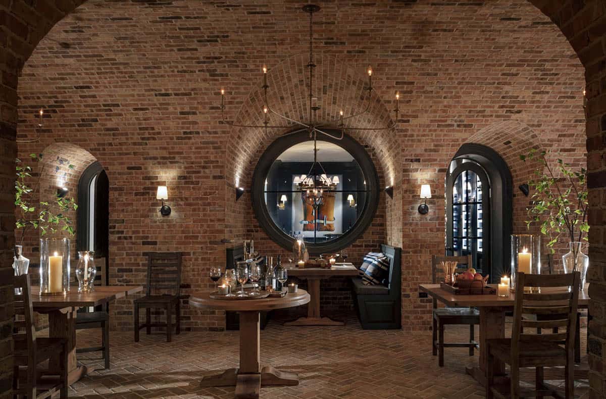 Wine cellar tasting room with brick arched ceiling, round porthole window, wood dining tables with candles, and arched wine storage niches
