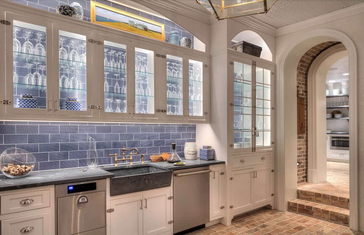 Butler's pantry with blue subway tile backsplash, glass-front cabinets displaying stemware, stone sink, brass faucet, and arch to brick cellar beyond