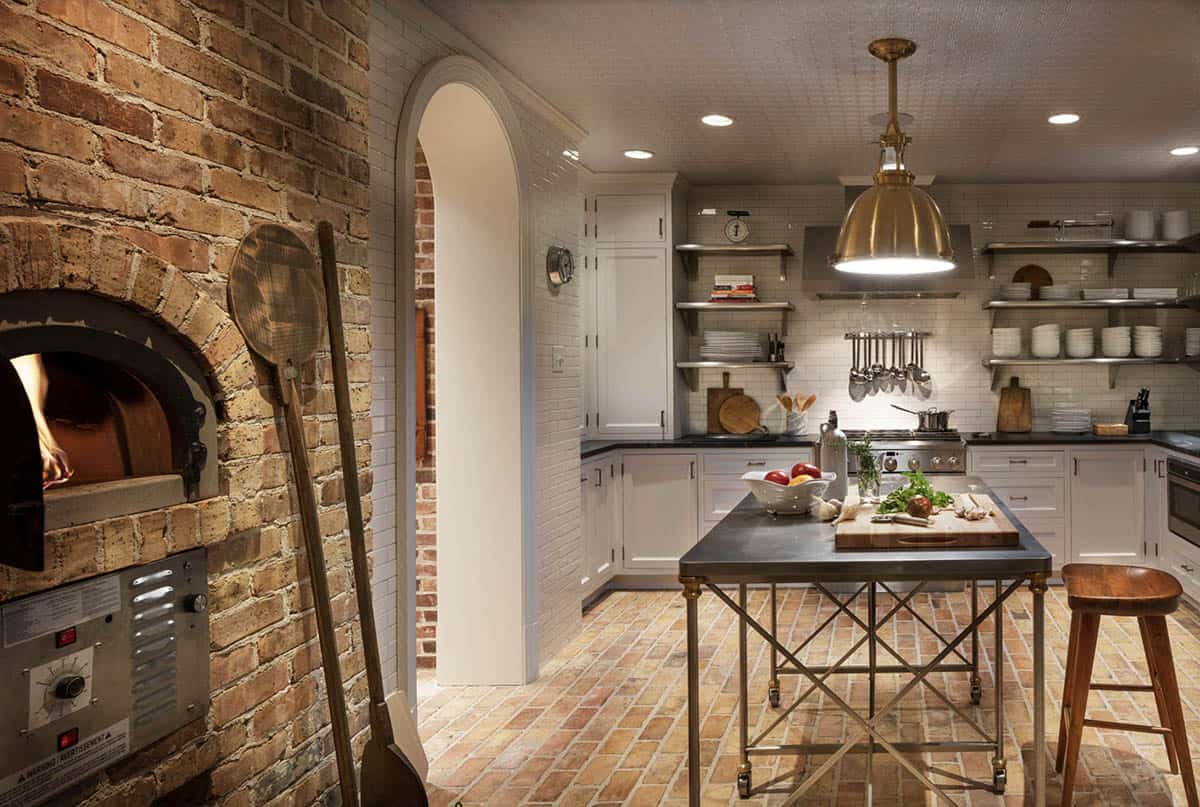 Lower-level catering kitchen with brick wood-fired pizza oven, white cabinetry, open metal shelving, brass pendant, and rolling prep island