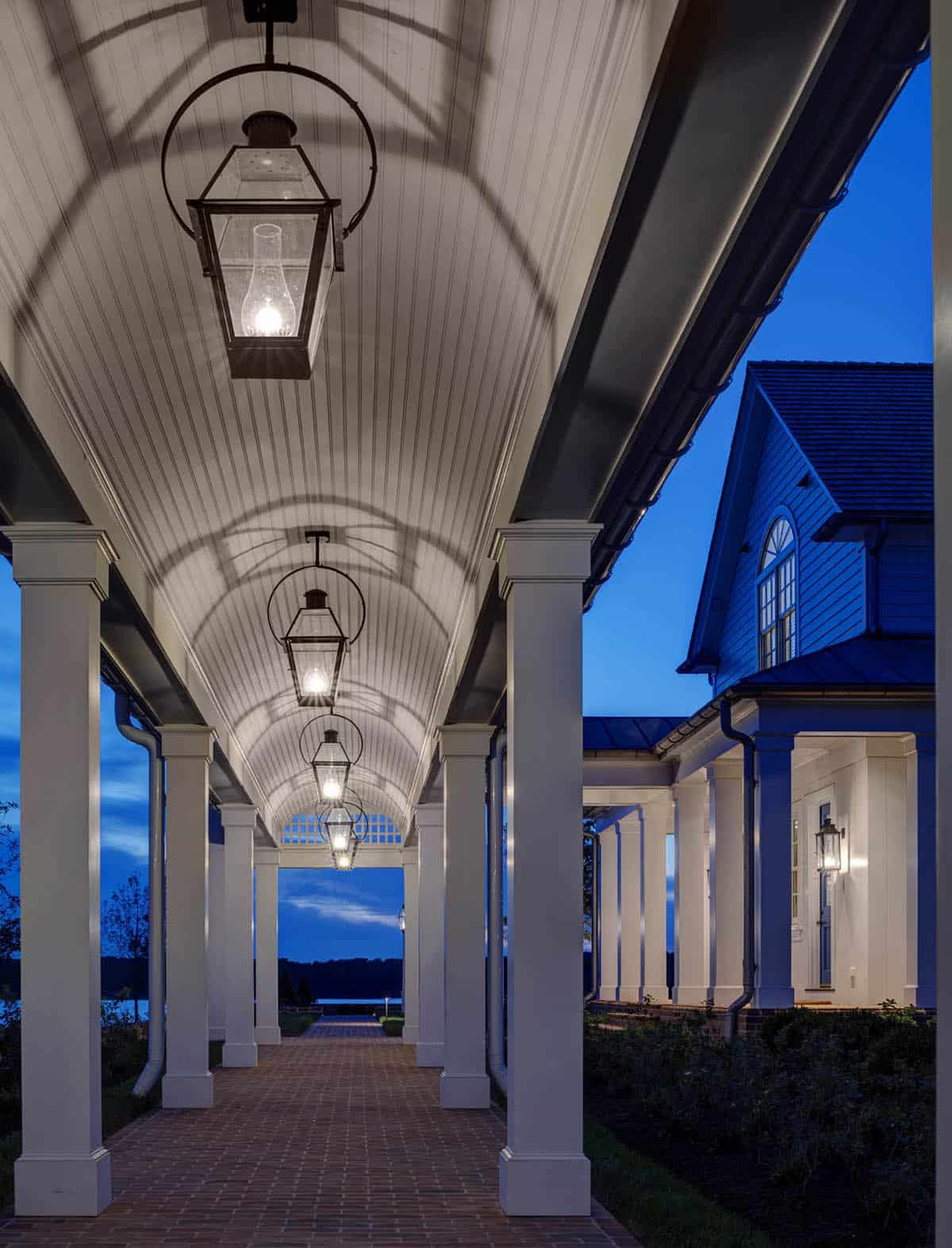 Illuminated covered walkway at dusk with repeating iron lanterns, barrel-vaulted beadboard ceiling, white columns, and view toward the river