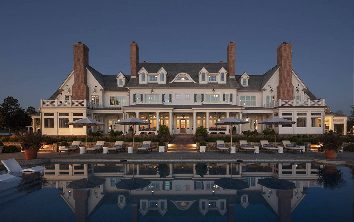 Rear facade of manor house at twilight perfectly reflected in the swimming pool, with lit colonnaded porch and four brick chimneys