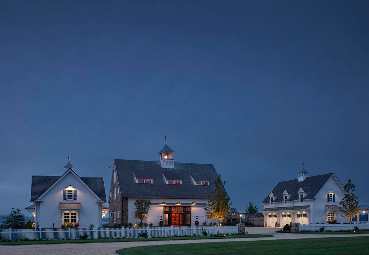 Twilight view of illuminated carriage house, stone entertainment barn, and garage outbuilding complex against a deep blue sky