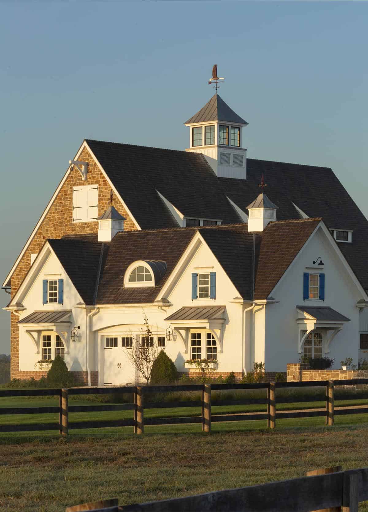 White carriage house at golden hour with cupola weather vane, blue shutters, arched dormer, and split-rail paddock fencing