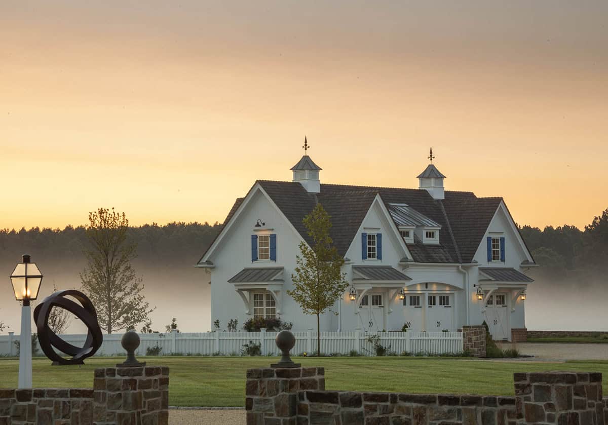 White carriage house at foggy sunrise with dual cupolas, blue shutters, misty river beyond, and circular metal sculpture on the lawn