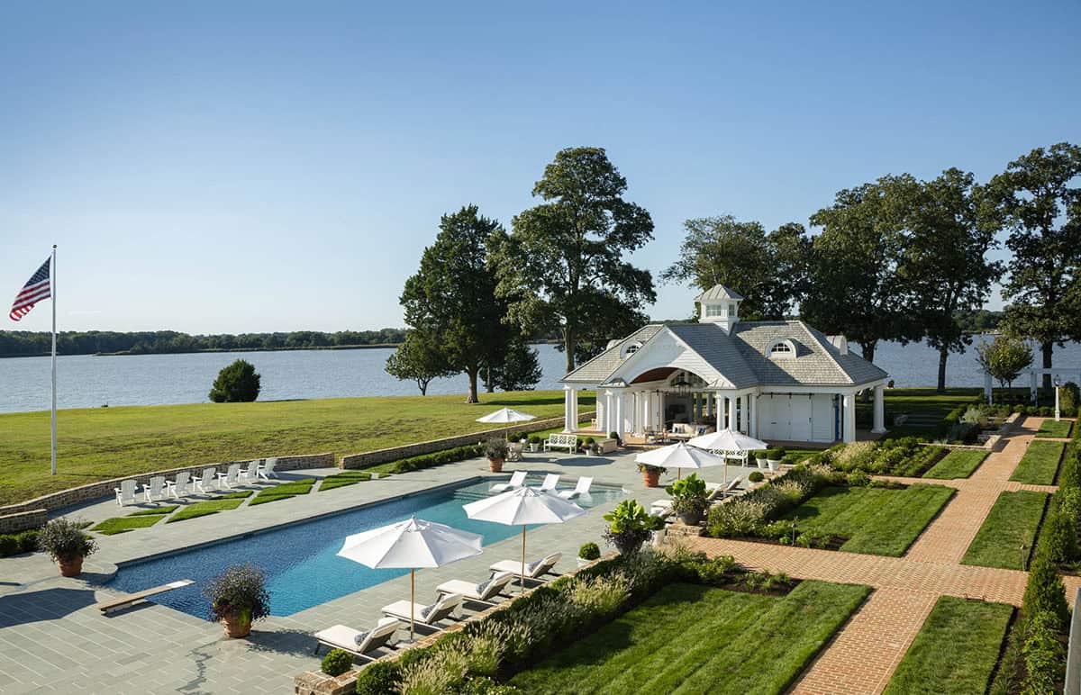 Aerial view of rectangular swimming pool, white pool house with cupola, formal garden beds, and American flag overlooking the river
