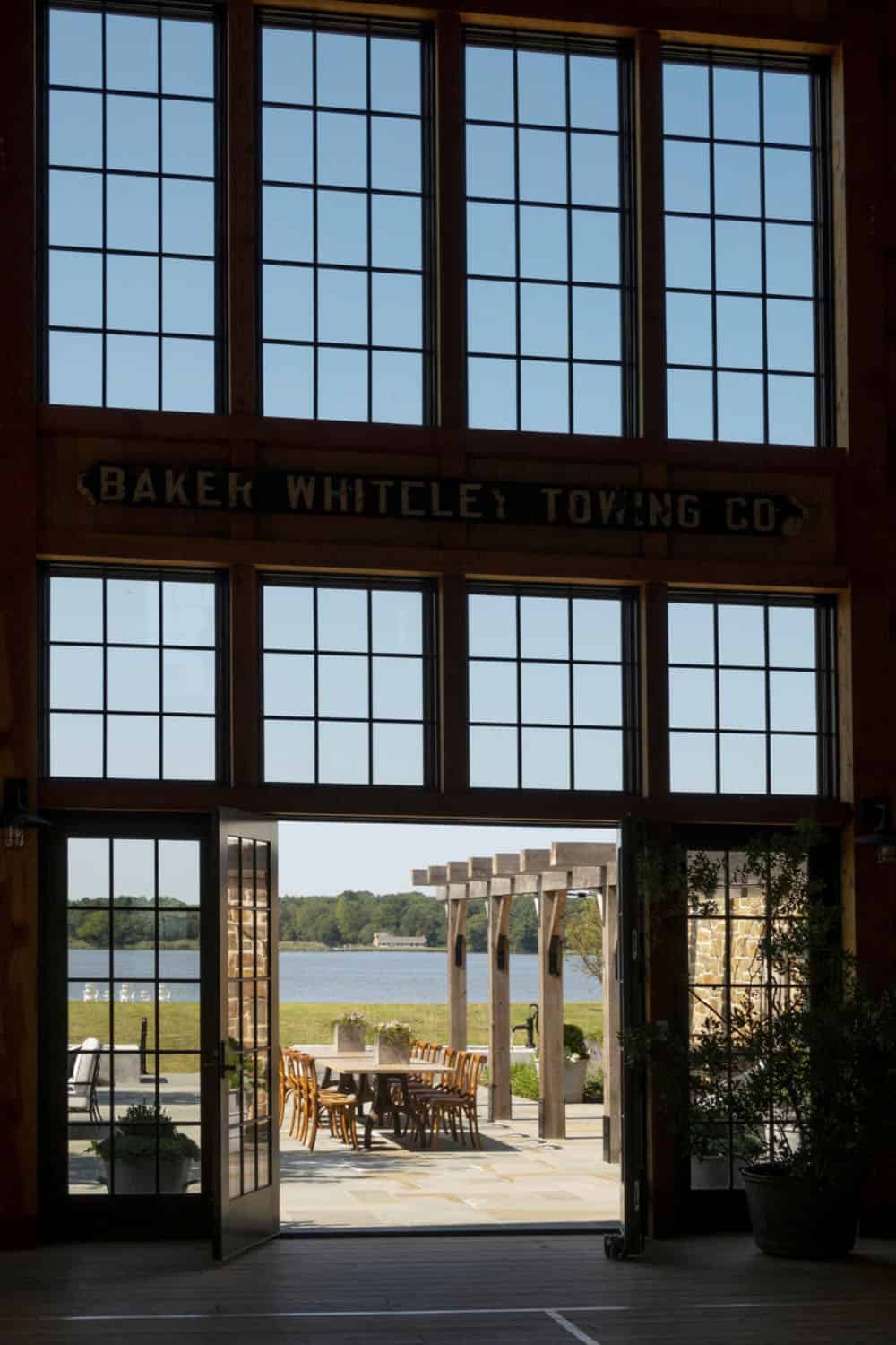 View through tall barn windows toward outdoor pergola dining terrace and river beyond, with antique towing company sign above