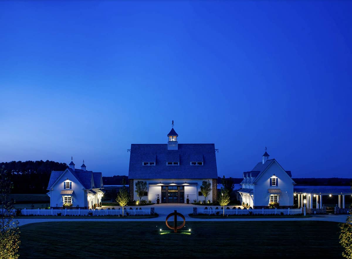 Front elevation of stone and white barn complex illuminated at night with cupola, weather vane, and circular sculpture focal point
