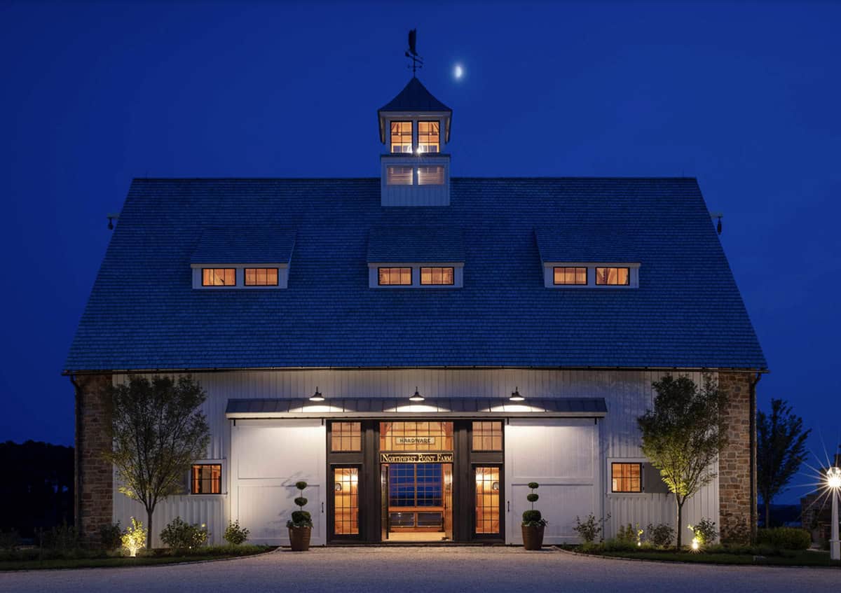 Northwest Point Farm barn at twilight with lit cupola, dormer windows glowing amber, stone flanking walls, and crescent moon above