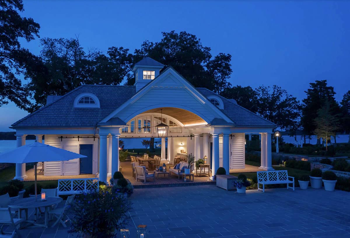Pool house at dusk with glowing open colonnade, teak furniture, lanterns, blue door, and reflections on the surrounding terrace