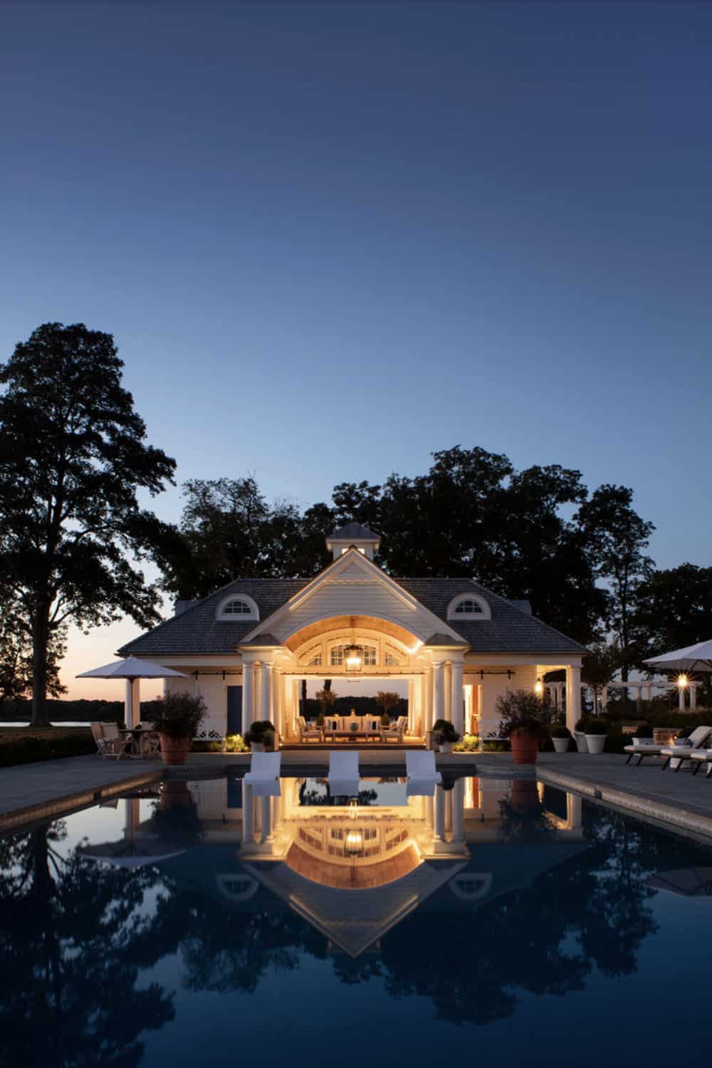 Pool house perfectly reflected in still swimming pool at twilight, with arched open pavilion glowing warmly against a blue dusk sky