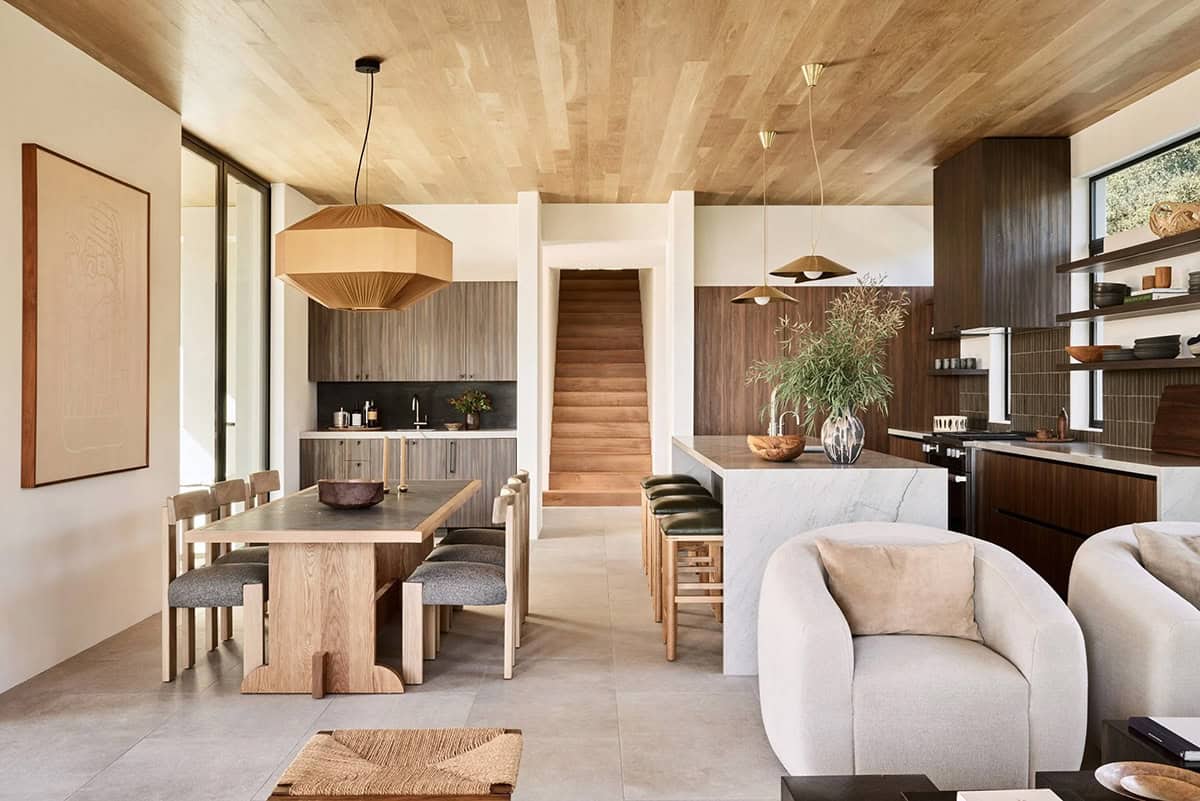 Open plan dining and kitchen with wood ceiling, oak dining table, marble island, and brass pendants