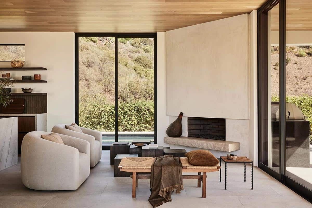 Living room with plaster fireplace, cream boucle sofas, woven bench, and floor-to-ceiling hillside views