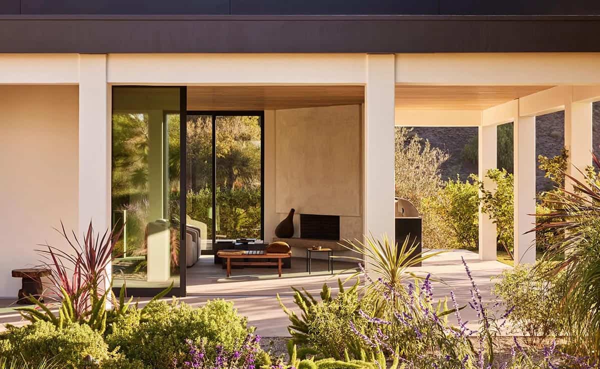 Modern home exterior with flat roof, open loggia, plaster fireplace visible through glass, and native garden