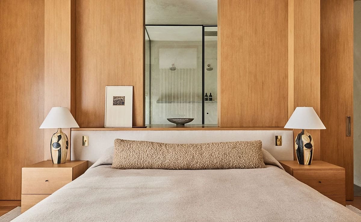 Primary bedroom headboard wall with integrated wood paneling, ceramic lamps, and peek-through shower beyond