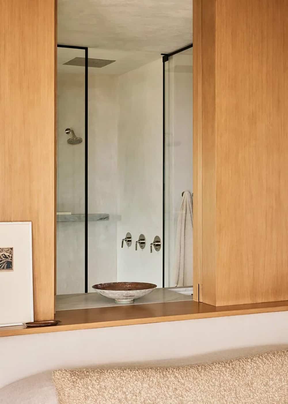 Wood-paneled bedroom wall with glass window view into primary shower with marble shelf