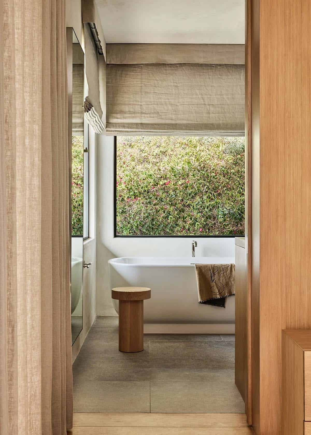 Primary bathroom with freestanding soaking tub, black-framed window, wood stool, and garden hedge view