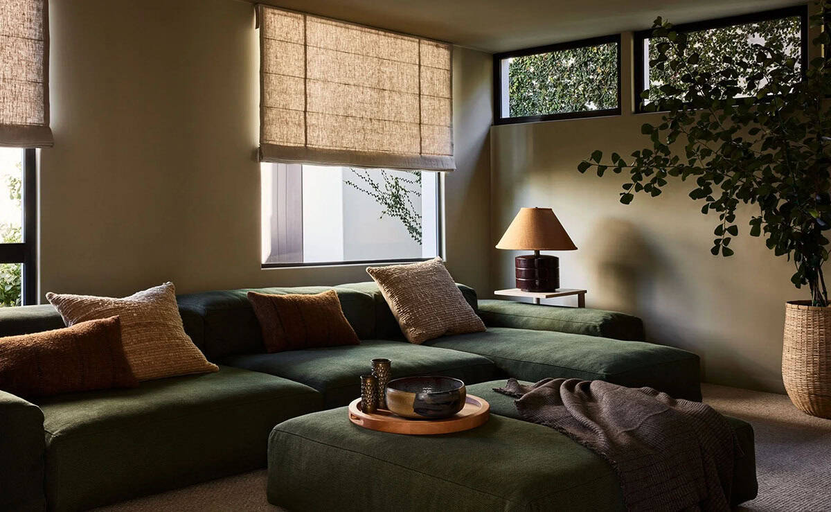 Moody media room with deep green sectional sofa, linen roman shades, table lamp, and potted fig tree