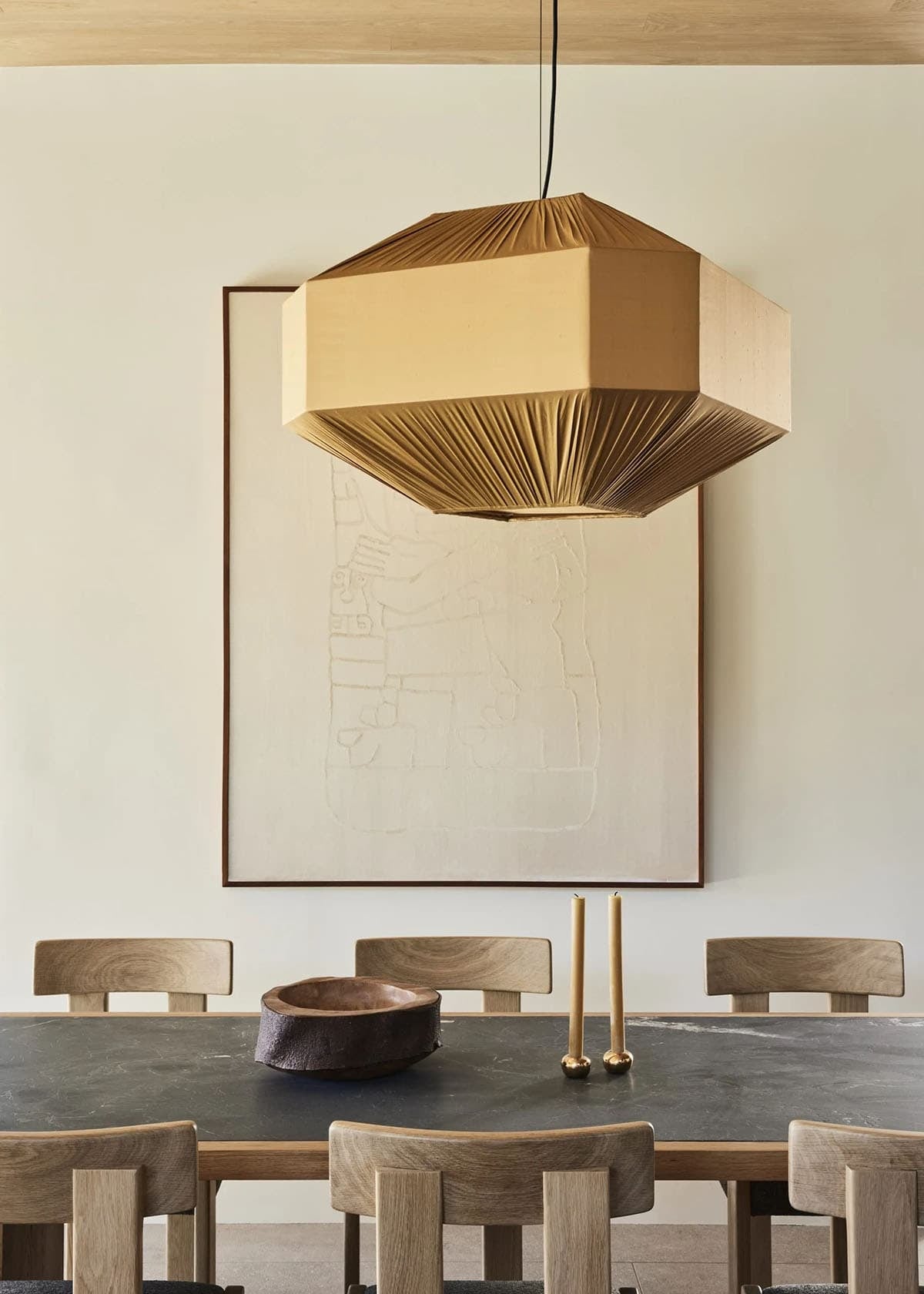 Close-up of oversized pleated pendant light above dining table with wood bowl, brass candlesticks, and large framed artwork