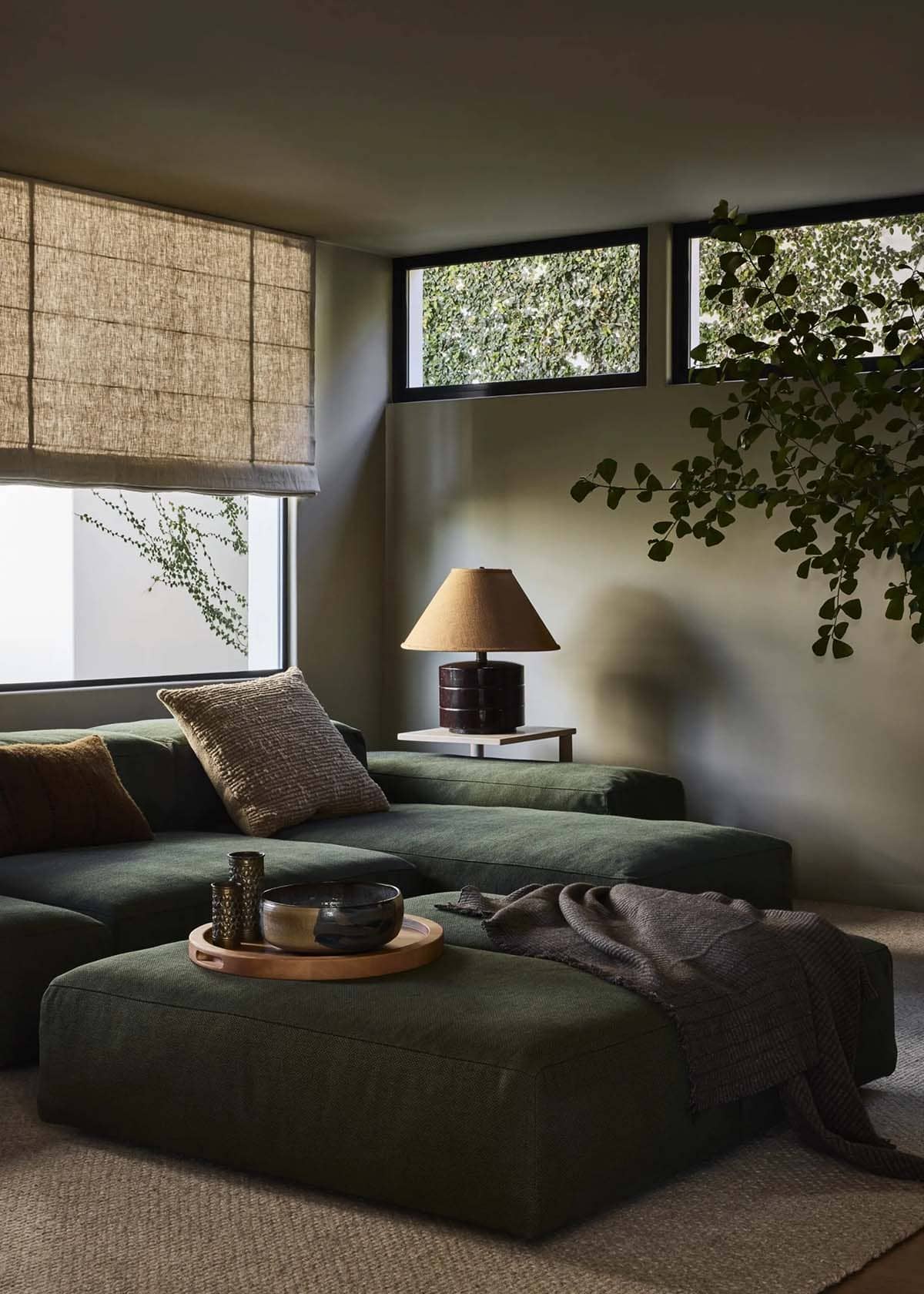 Moody corner of media room with deep green sectional, wood tray ottoman, table lamp, and trailing vine plant