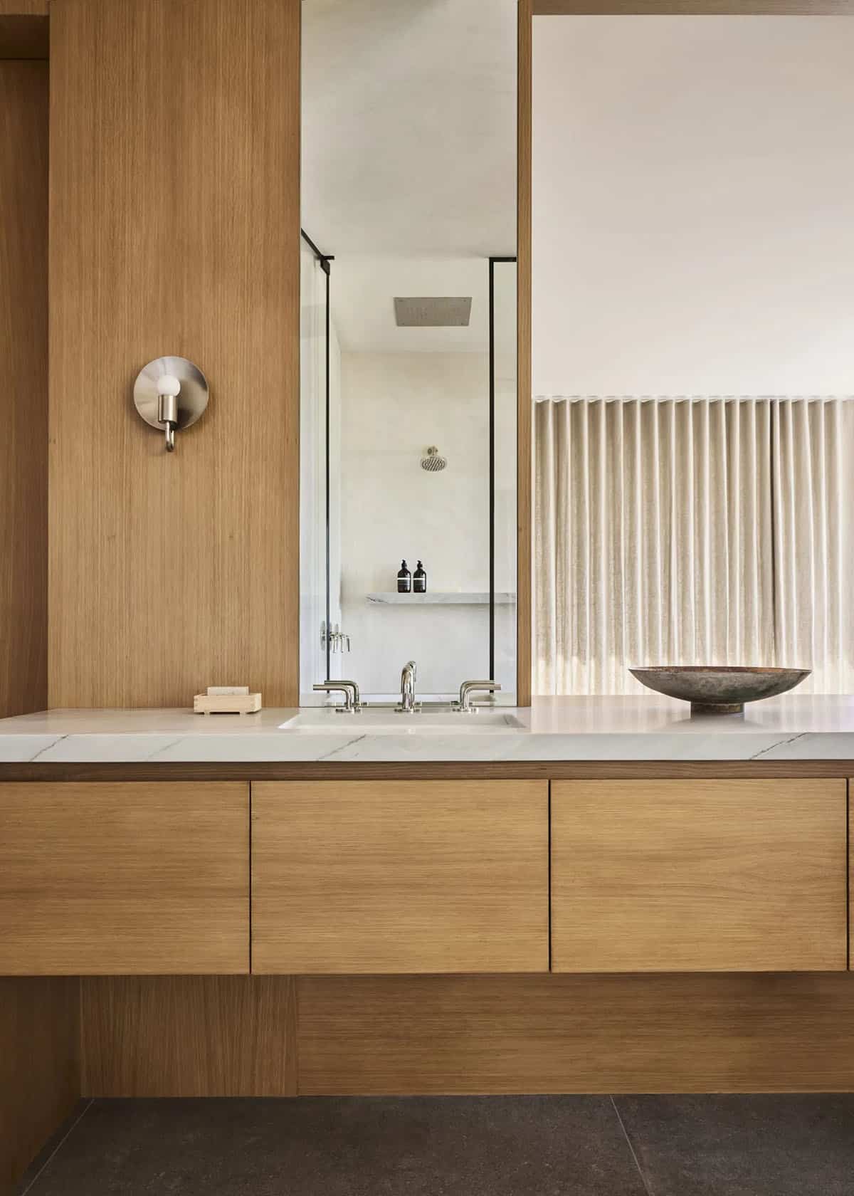 Primary bathroom vanity close-up with oak cabinetry, marble countertop, mirror reflecting shower, and linen drapes