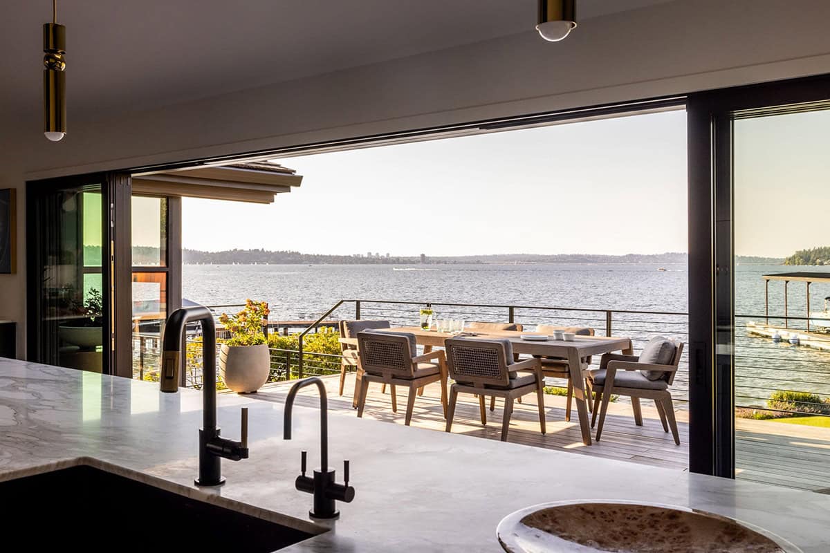 Kitchen with marble island and black faucets opening to waterfront deck with lake views through bifold doors