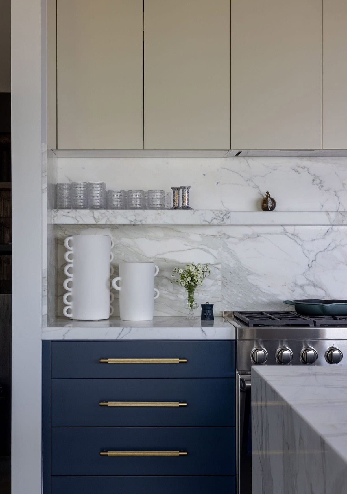 Kitchen detail with navy lower cabinets, brass hardware, marble backsplash, and open floating shelf
