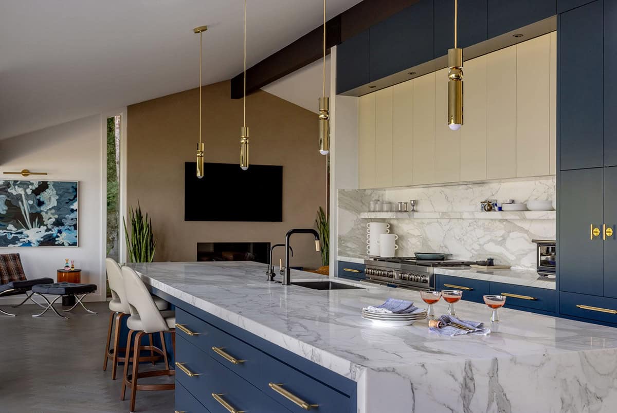 Open-plan kitchen with navy cabinets, marble waterfall island, brass pendant lights, and family room beyond