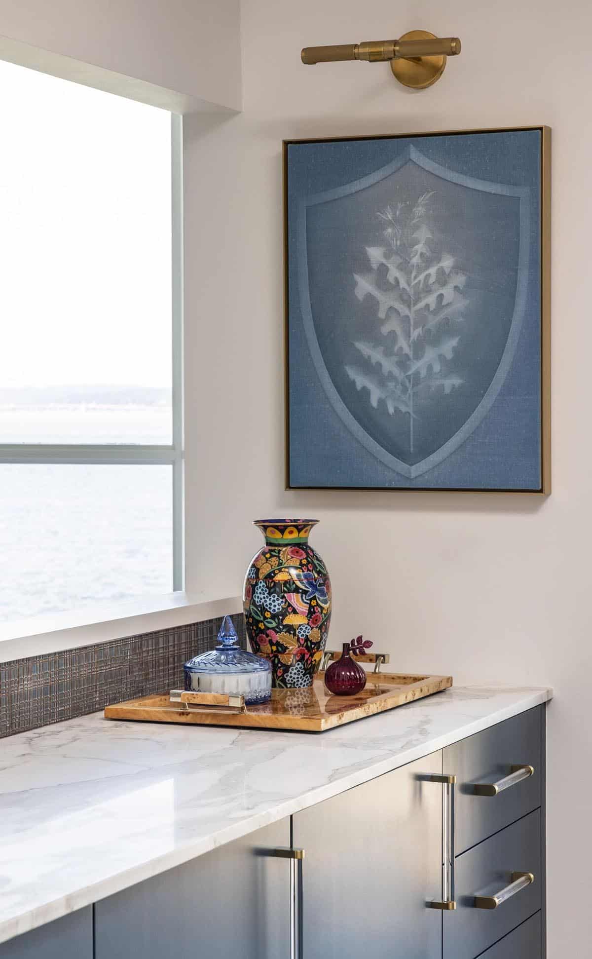 Kitchen vignette with marble countertop, colorful ceramic vase, botanical artwork, and lake window view