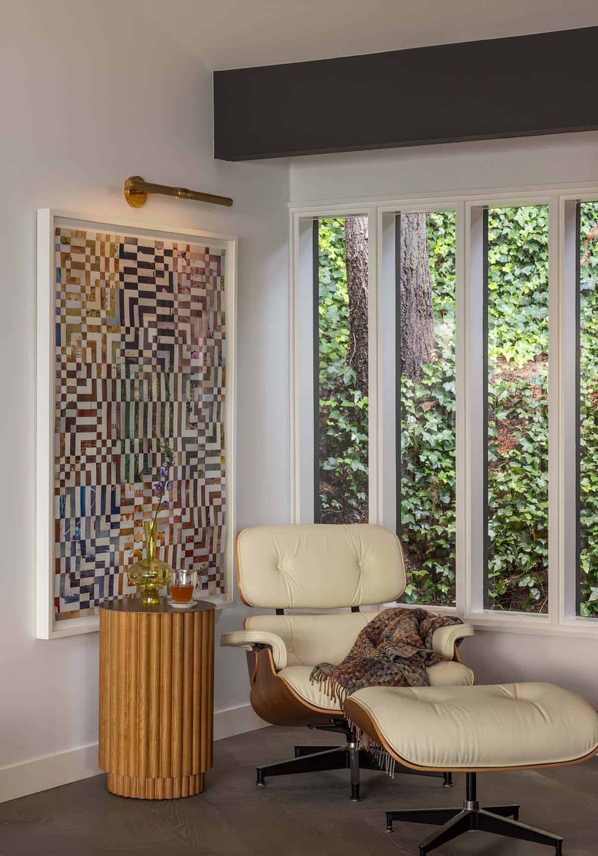 Reading nook with Eames lounge chair, fluted side table, pixelated artwork, brass sconce, and ivy-covered garden window