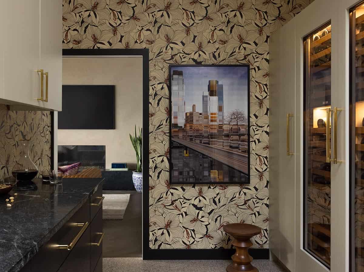 Wet bar with bold floral wallpaper, dark granite countertop, brass hardware, framed cityscape artwork, and wine fridge