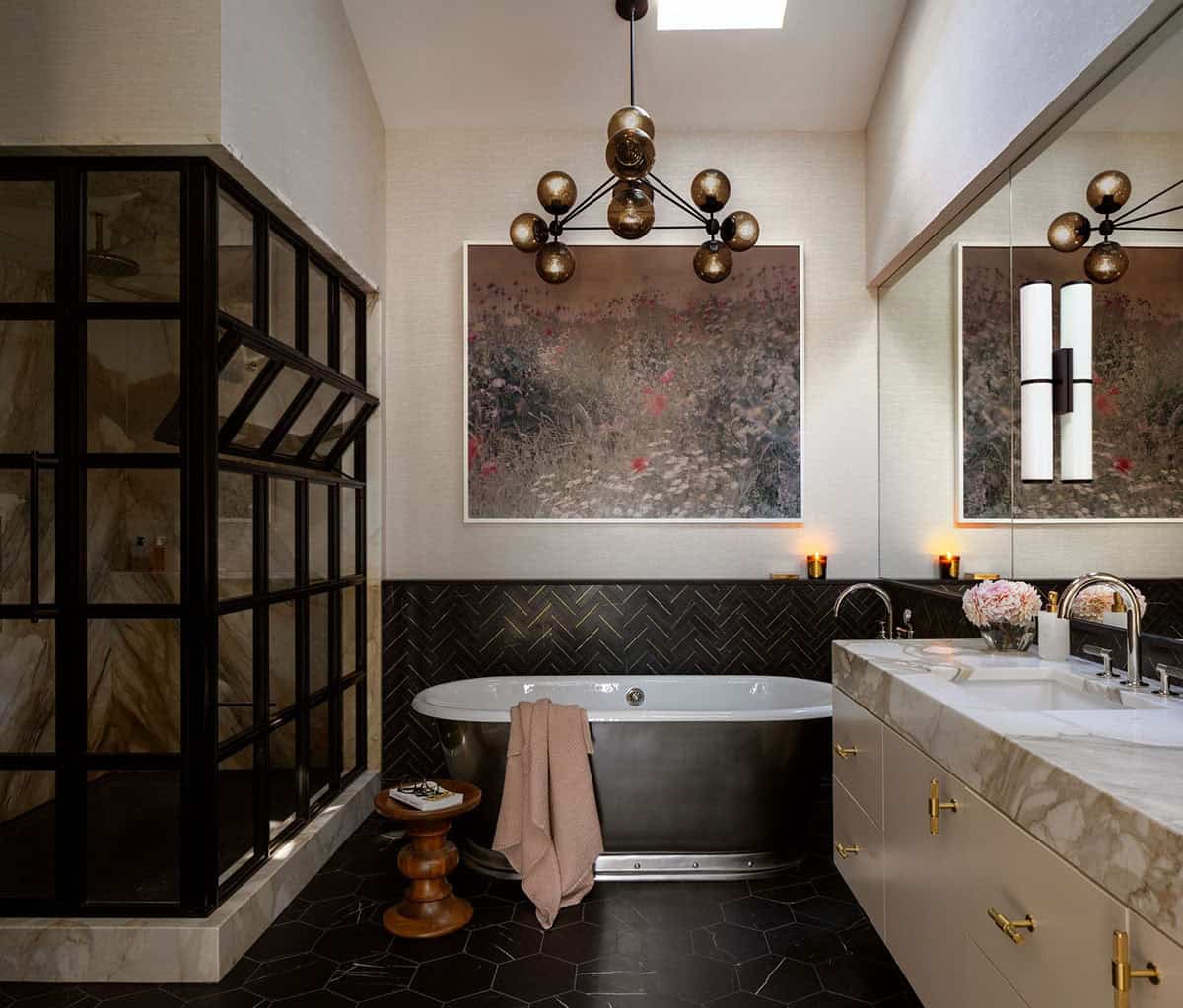 Primary bathroom with black freestanding tub, black herringbone tile, steel-framed shower, gold globe chandelier, and floral artwork