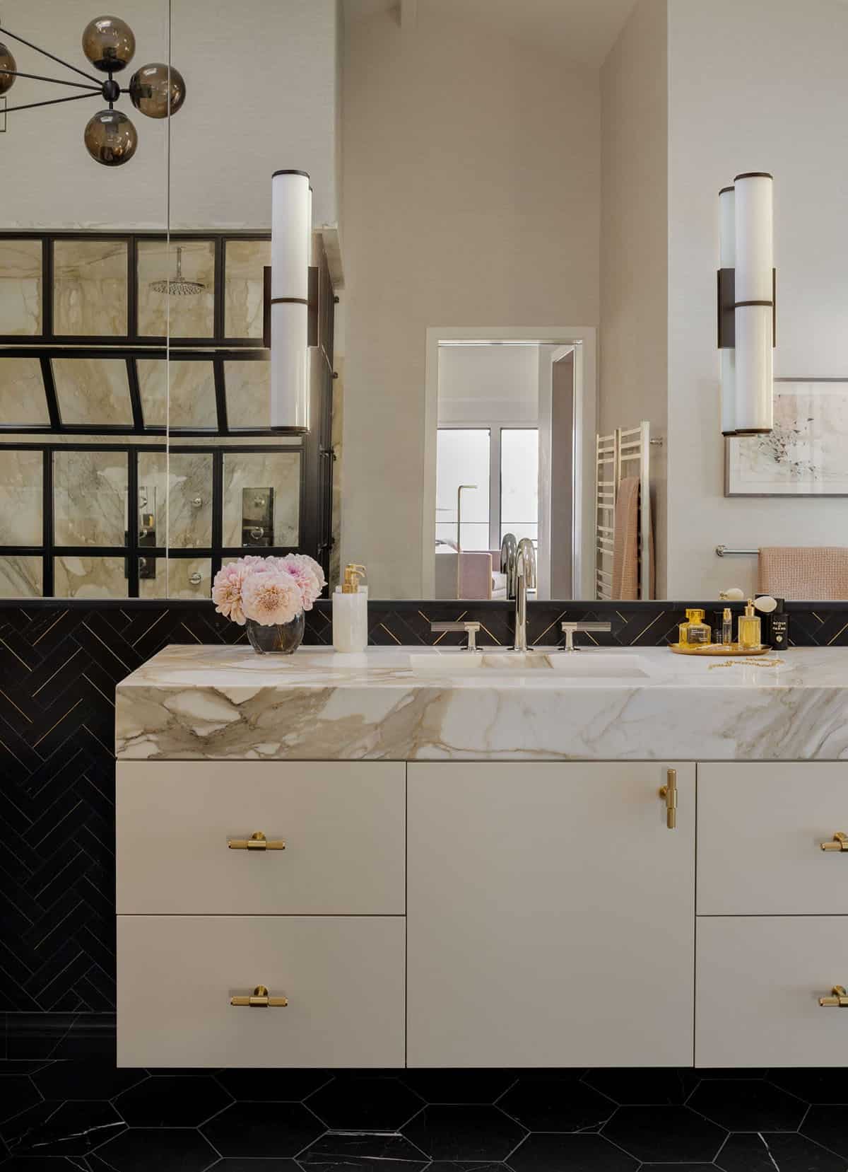 Primary bathroom vanity with cream cabinets, marble countertop, brass hardware, and cylinder wall sconces