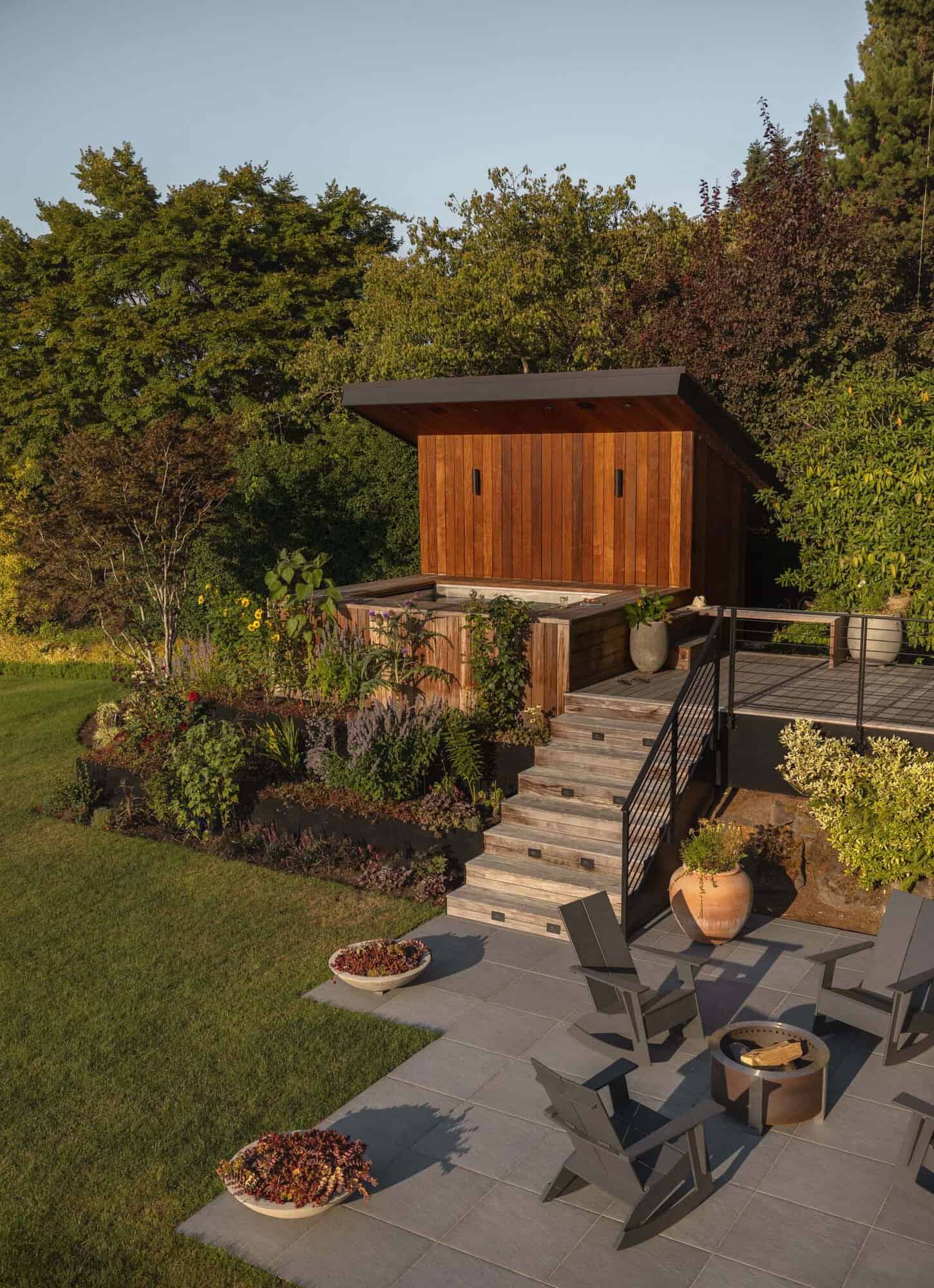 Backyard with cedar sauna, hot tub, fire pit patio, Adirondack chairs, and terraced garden beds at golden hour