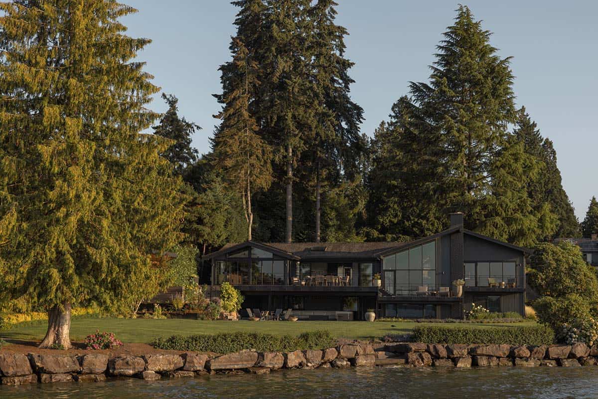 Mid-century modern waterfront home viewed from the lake with sweeping lawn, stone seawall, and tall evergreens on Mercer Island