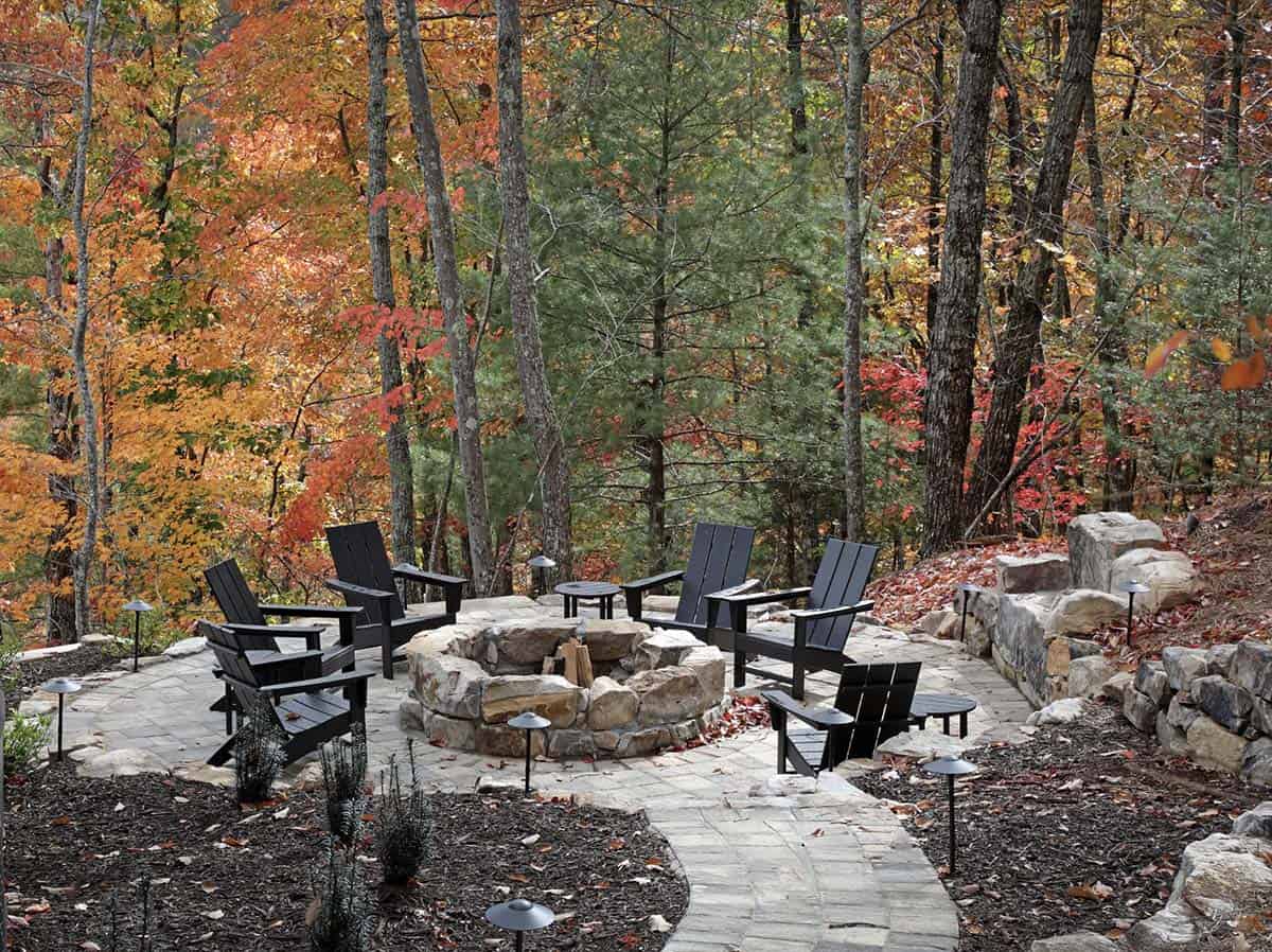 Stone fire pit surrounded by modern black Adirondack chairs amid fall forest