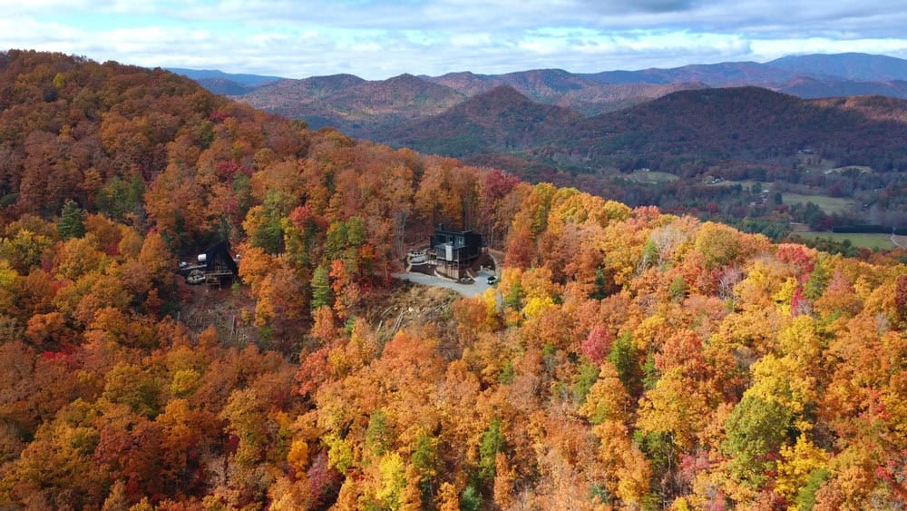 Aerial view of dark modern home perched on forested mountain ridge with valley beyond