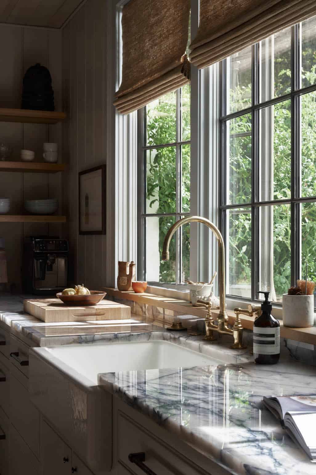 Farmhouse kitchen sink with brass bridge faucet, marble countertops, and steel-framed garden window