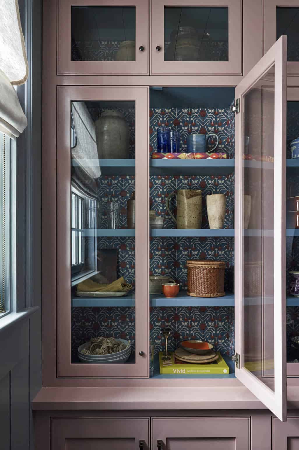 Open glass-front cabinet with pink exterior, blue patterned wallpaper lining, and collected ceramics