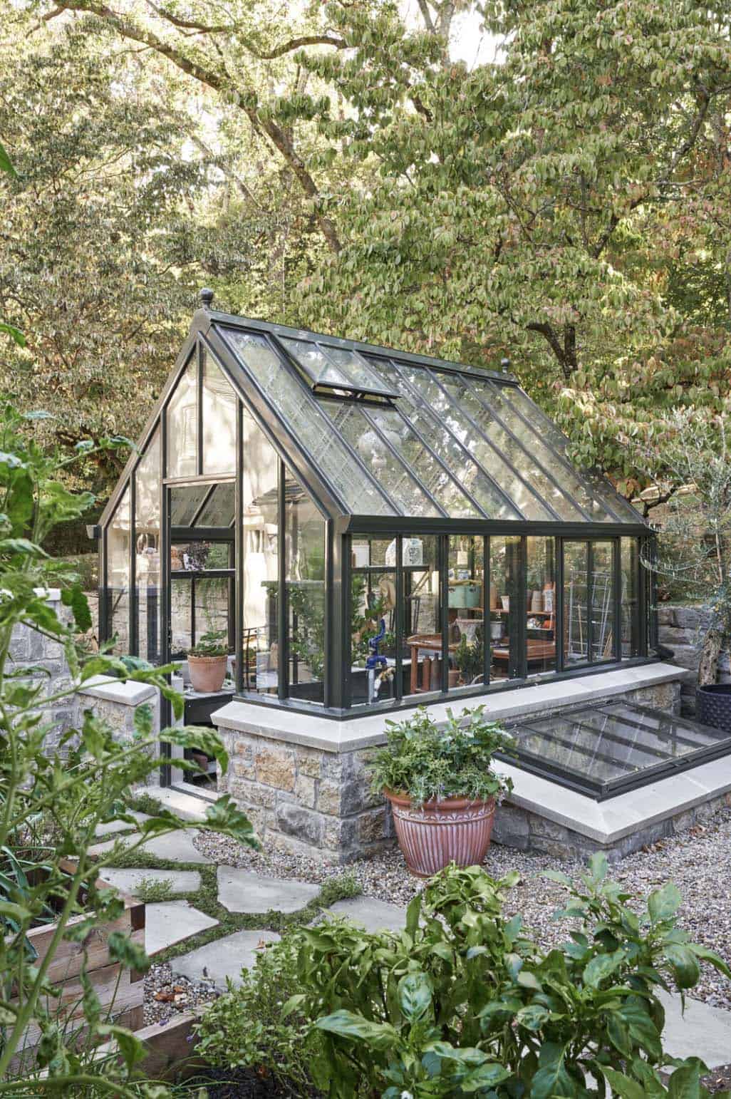 Victorian-style glass greenhouse with black steel frame on stone base surrounded by garden plantings