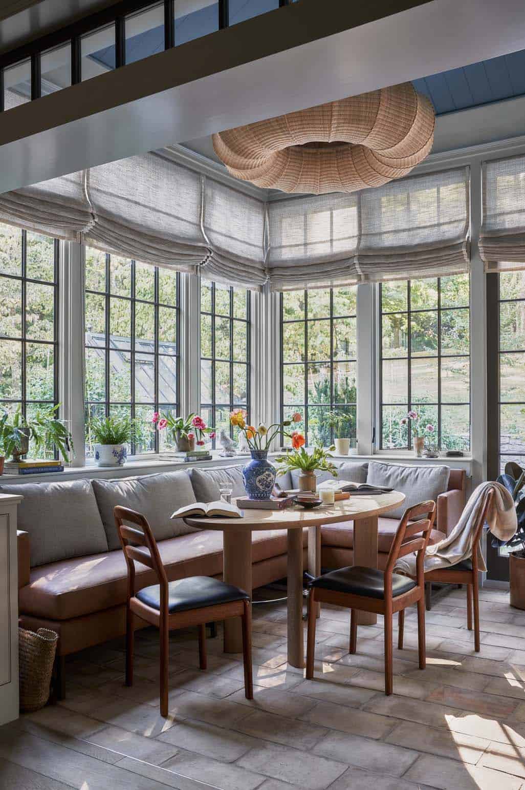 Sunroom breakfast nook with curved banquette, round table, rattan pendant, and steel-framed windows