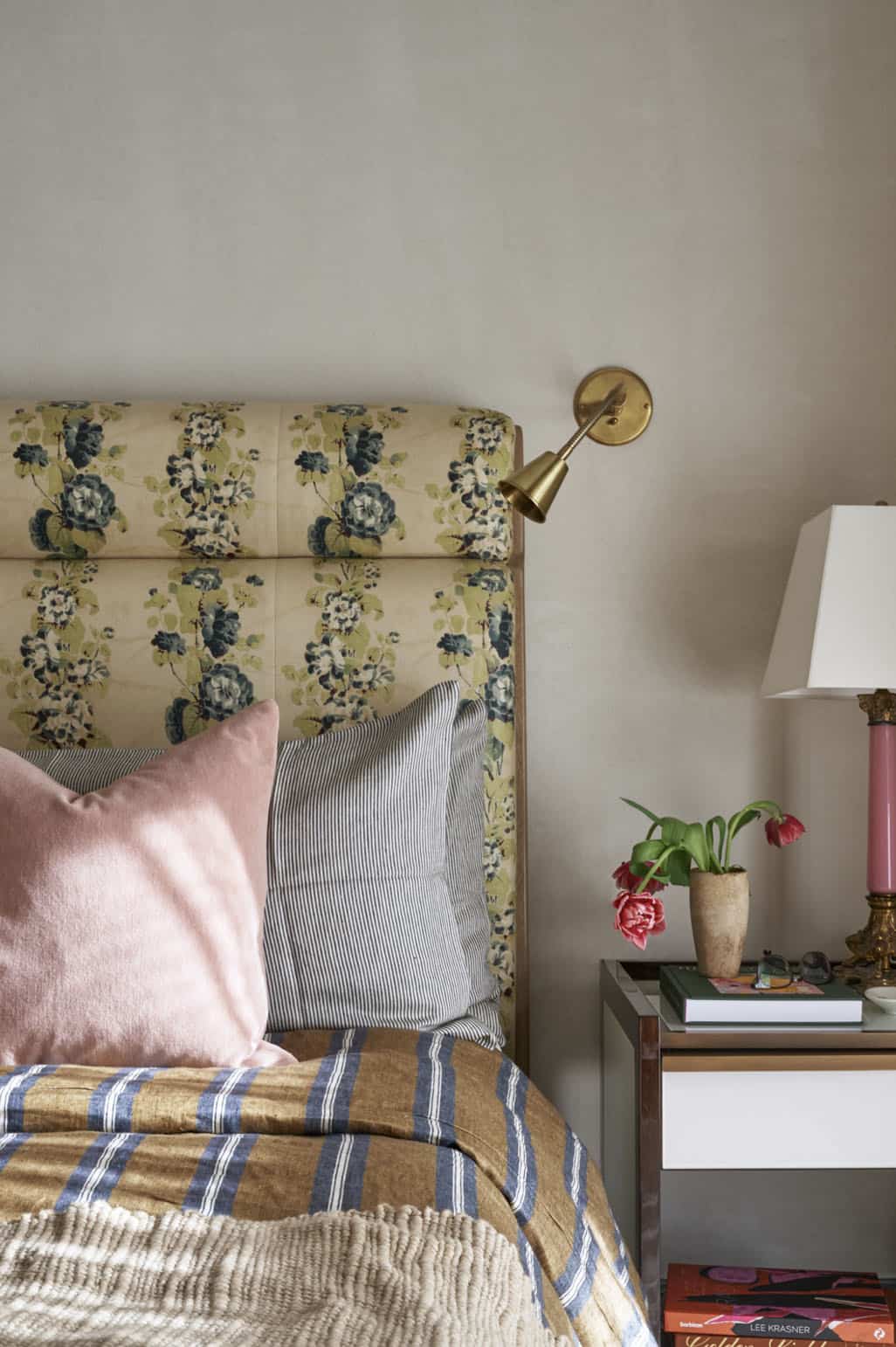 Floral upholstered headboard with pink and striped pillows, brass wall sconce, and pink table lamp