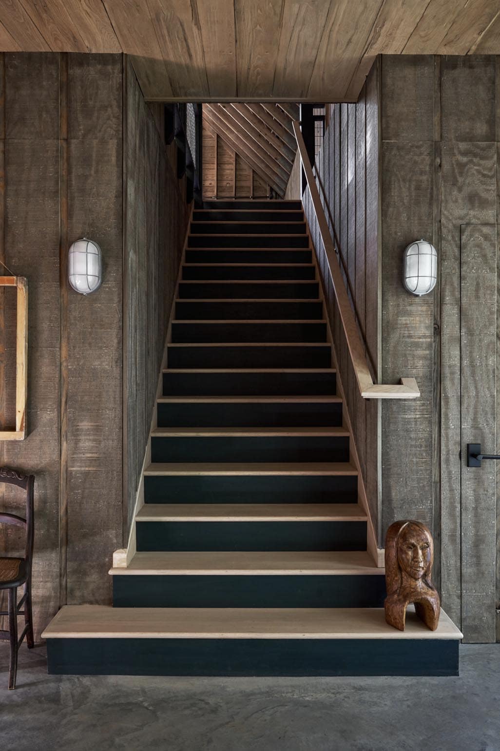 Steep staircase with black-painted risers, raw wood treads, industrial bulkhead sconces, and weathered board walls