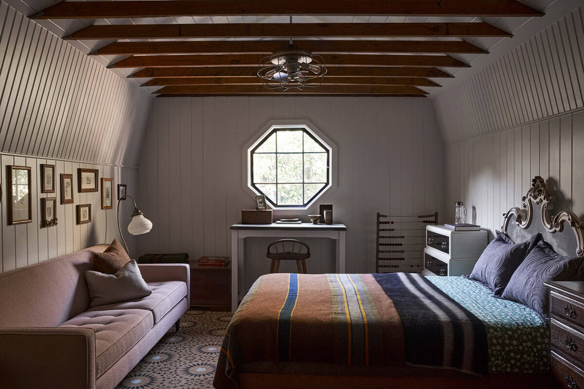 Attic bedroom with gambrel ceiling, exposed beams, octagonal window, ornate headboard, and striped blanket